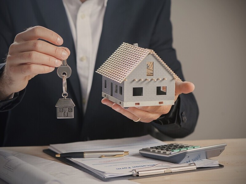 Person holding house model and keys above insurance documents on a desk
