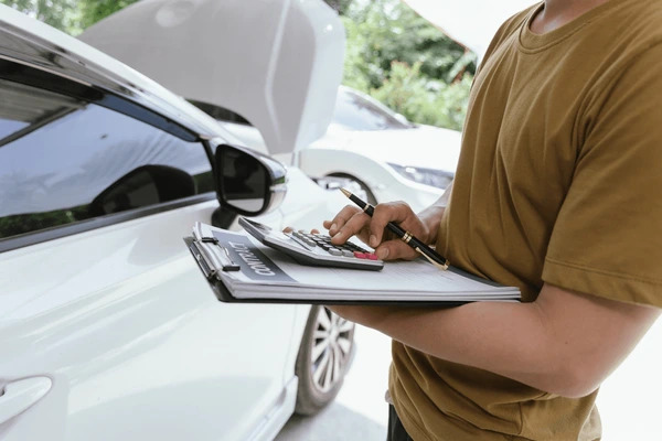 Person holding a clipboard and calculator beside a car with the hood open
