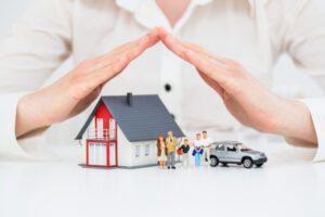 Hands protecting a model house, family, and car symbolizing insurance coverage