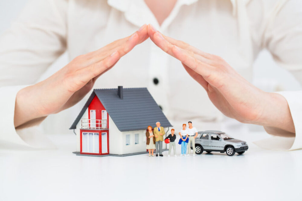 Hands protecting a model house, family, and car symbolizing insurance coverage